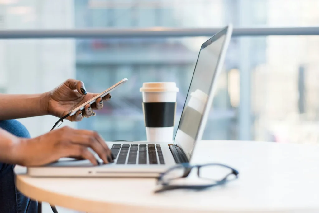 Close-up of a woman multitasking at a table with her left hand typing on a laptop and her right hand holding a smartphone, with a blurred cityscape background.