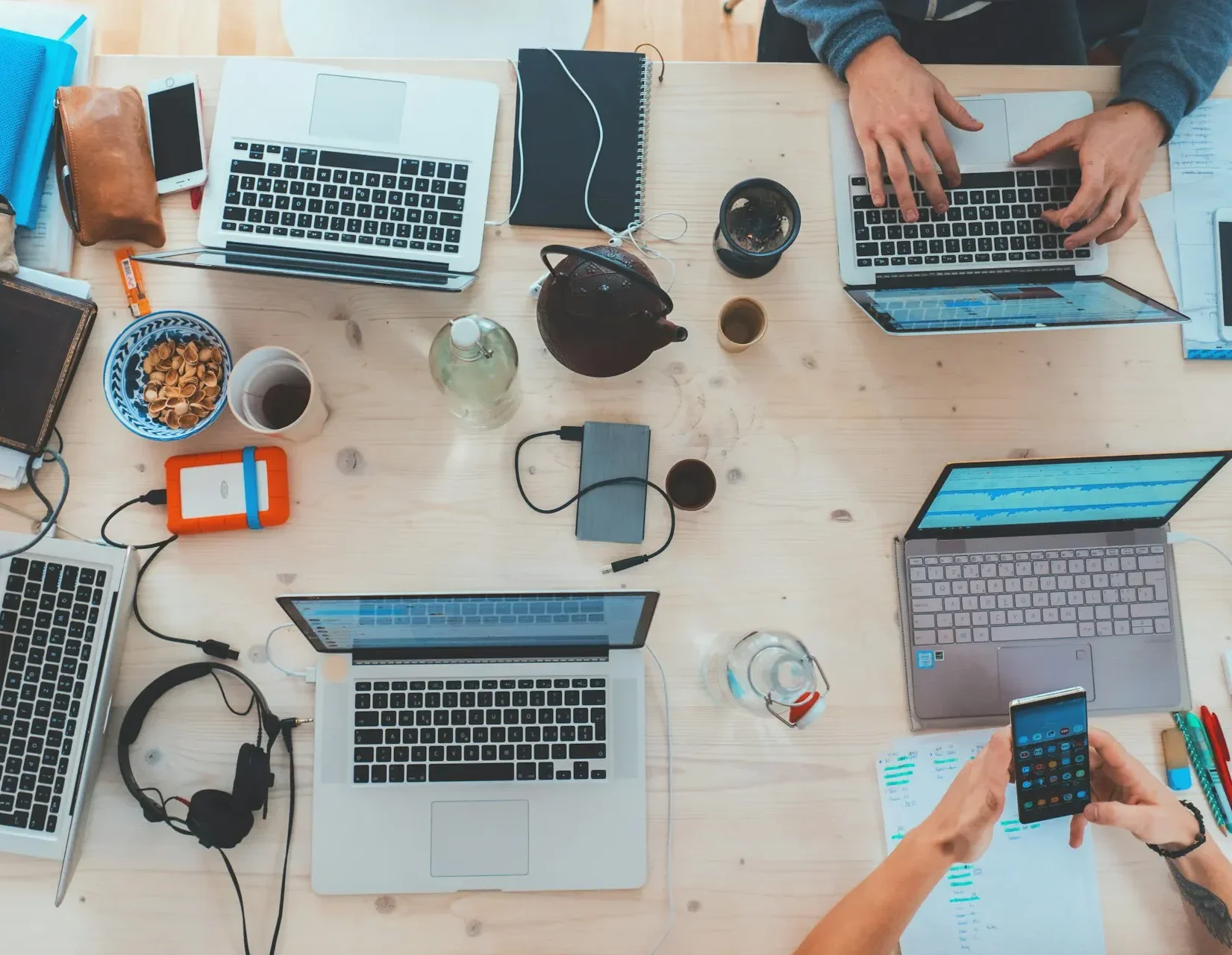 Top-down view of four laptops on a table with hands working on them, accompanied by coffee, snacks, and computer accessories.