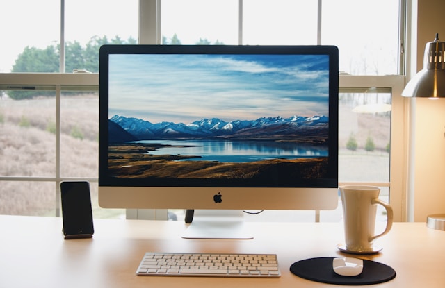 Modern iMac desktop setup with outdoor landscape on screen, accompanied by iPhone, keyboard, mouse and coffee cup on a clean wooden desk near a window.