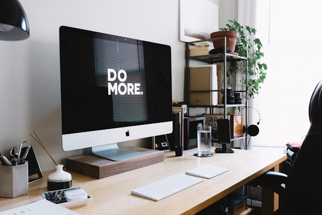 Minimalist home office with an iMac displaying 'DO MORE' on the screen, desk supplies, and shelving in the background.